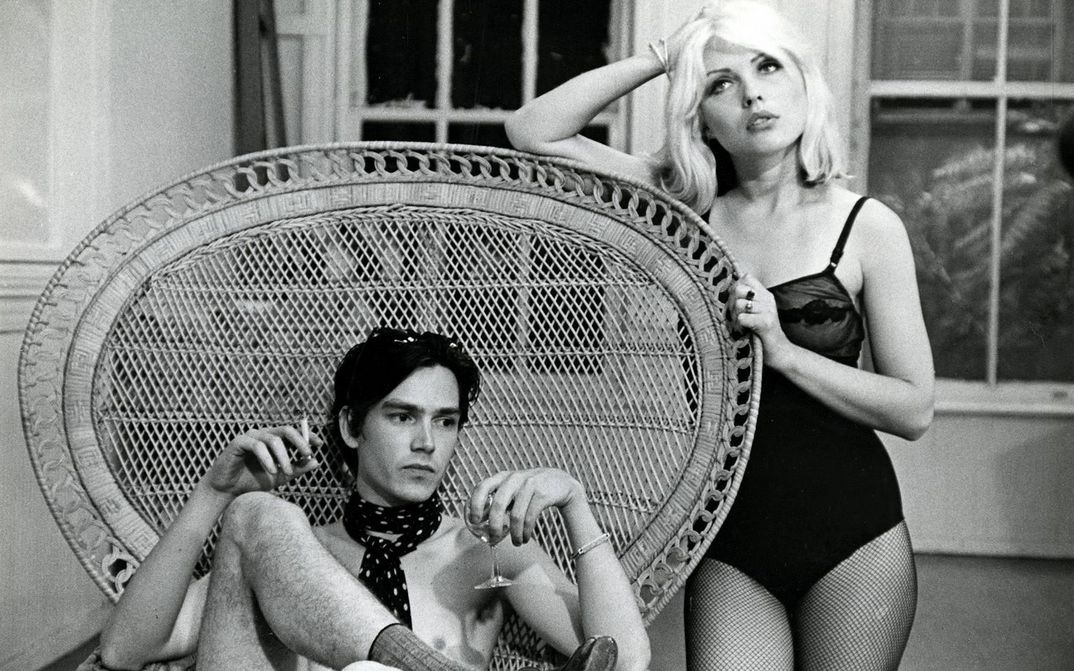 Filmstill from UNMADE BEDS: A man is sitting in a wicker chair with a very large round backrest. He is smoking and drinking from a champagne glass. A woman in a bodysuit and fishnet tights is leaning on the backrest with her elbow.