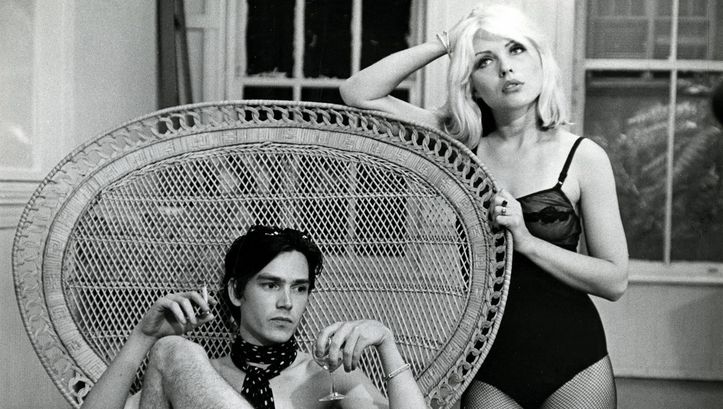 Filmstill from UNMADE BEDS: A man is sitting in a wicker chair with a very large round backrest. He is smoking and drinking from a champagne glass. A woman in a bodysuit and fishnet tights is leaning on the backrest with her elbow.