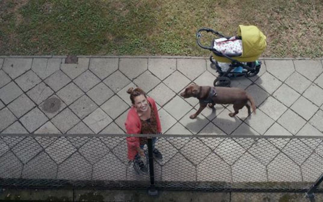 Film still from THE BALCONY MOVIE. View from a balcony on the street, a woman with a dog and stroller looks up.