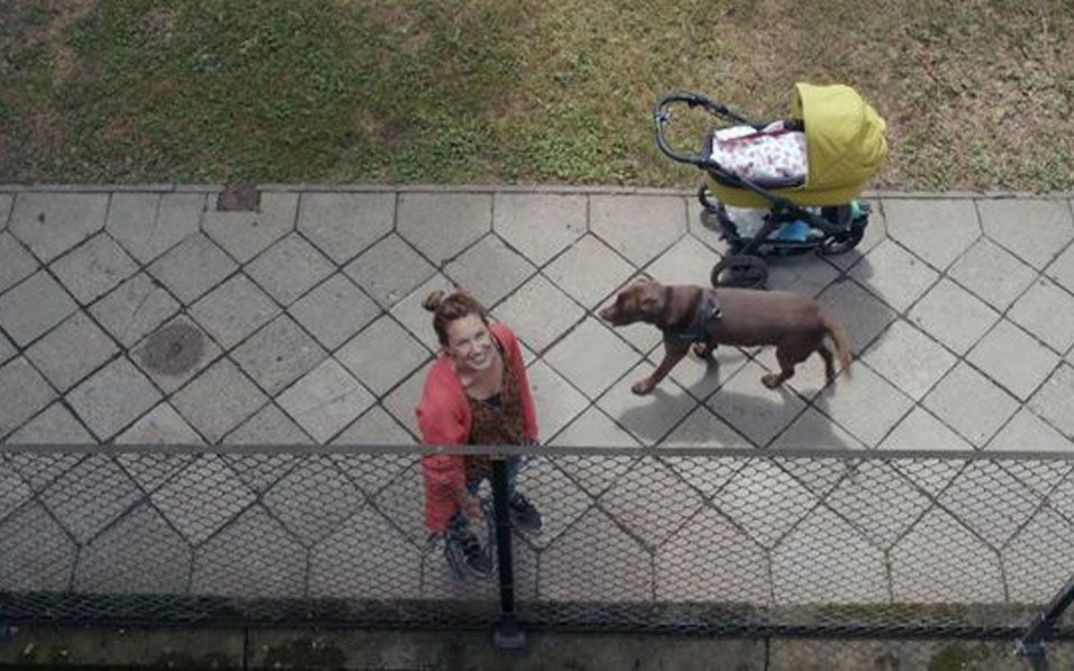 Film still from THE BALCONY MOVIE. View from a balcony on the street, a woman with a dog and stroller looks up.