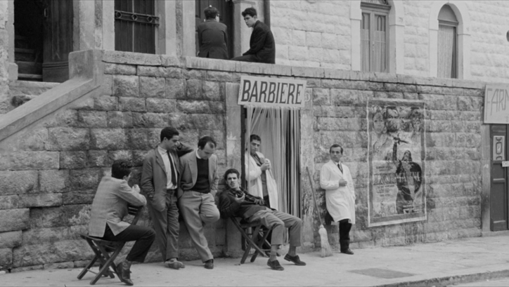 Film still from I BASILISCHI: A group of men are sitting and standing bored in front of a barber shop.