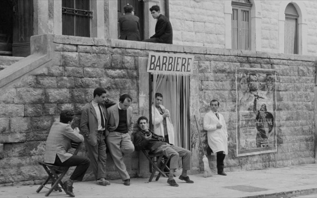 Film still from I BASILISCHI: A group of men are sitting and standing bored in front of a barber shop.