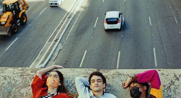 Still from the film "Três tigres tristes" by Gustavo Vinagre. We see the shoulders and heads of three young people laying down on a concrete bridge, while traffic flows beneath them.