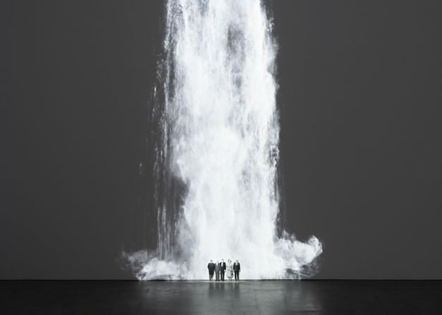 Installation view of Walid Raad’s „Comrade leader, comrade leader, how nice to see you“. A waterfall projected unto a wall; at its foot five figures standing next to each other.