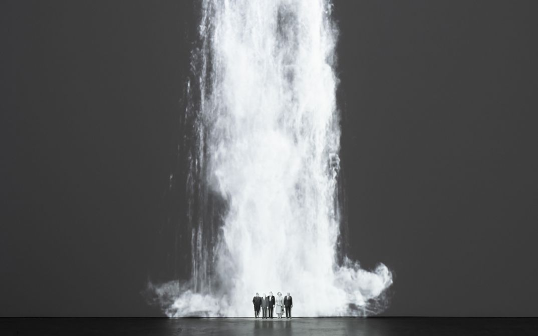 Installation view of Walid Raad’s „Comrade leader, comrade leader, how nice to see you“. A waterfall projected unto a wall; at its foot five figures standing next to each other.