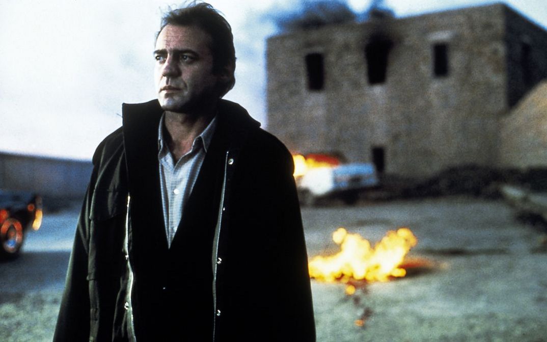 Filmstill aus DIE FÄLSCHUNG. Der Schauspieler Bruno Ganz im Vordergrund, im Hintergrund unscharf ein brennendes Haus.