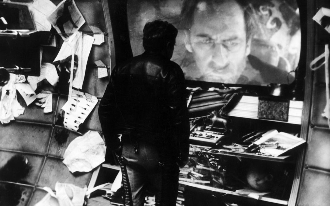 Film still from SOLARIS: In a room full of objects, one man looks at another on a large screen.