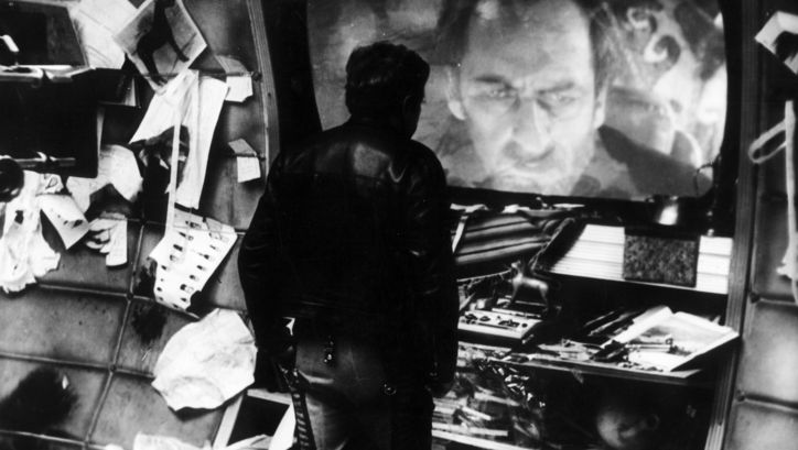 Film still from SOLARIS: In a room full of objects, one man looks at another on a large screen.