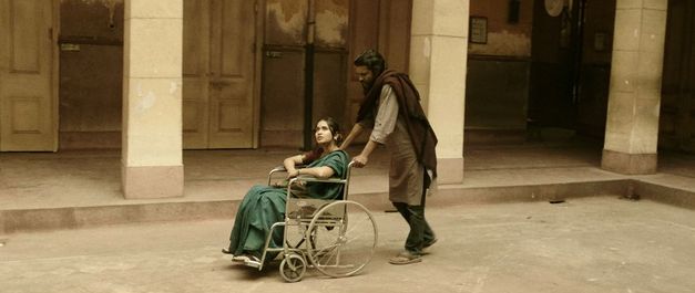 Filmstill aus dem Film "Those Who Do Not Drown" von Naeem Mohaiemen. Eine Frau sitzt in einem Rollstuhl und wird von einem Mann durch einen Hof geschoben.