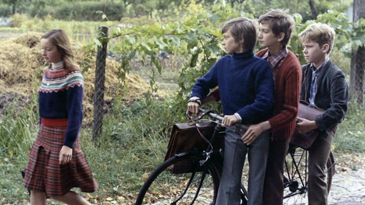 Filmstill aus MES PETITES AMOUREUSES: Ein Mädchen läuft mit einer Schultasche die Straße entlang. Drei Jungen, die an einem Fahrrad stehen, schauen ihr hinterher.
