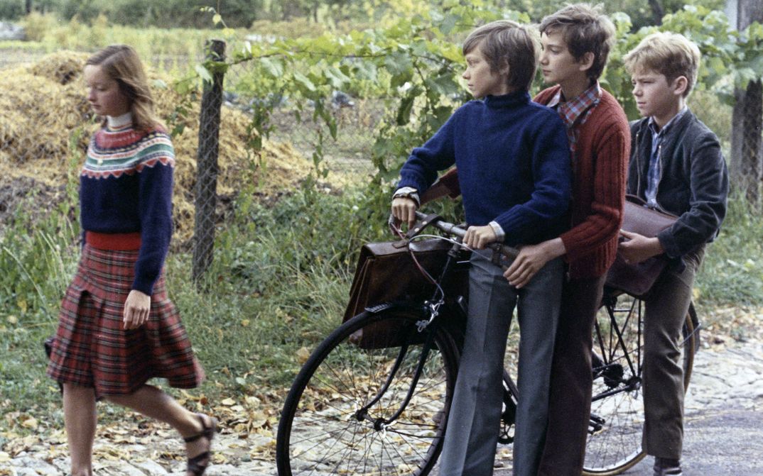 Filmstill aus MES PETITES AMOUREUSES: Ein Mädchen läuft mit einer Schultasche die Straße entlang. Drei Jungen, die an einem Fahrrad stehen, schauen ihr hinterher.