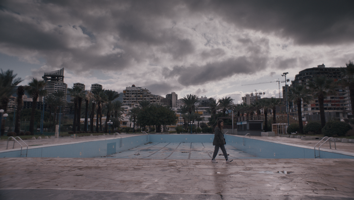 Filmstill aus THE SEA AHEAD. Panoramaaufnahme einer Stadtlandschaft mit Hochhäusern, einem leeren Schwimmbad und bewölktem Himmel.