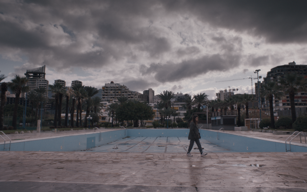 Filmstill aus THE SEA AHEAD. Panoramaaufnahme einer Stadtlandschaft mit Hochhäusern, einem leeren Schwimmbad und bewölktem Himmel.