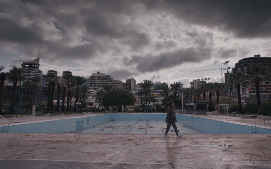 Filmstill aus THE SEA AHEAD. Panoramaaufnahme einer Stadtlandschaft mit Hochhäusern, einem leeren Schwimmbad und bewölktem Himmel.