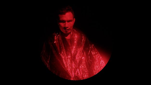 Filmstill aus dem Film "Yarokamena" von Andrés Jurado. Runder Bildausschnitt. Ein Mann mit einem silbernen Umhang wird von rotem Licht angeleuchtet.