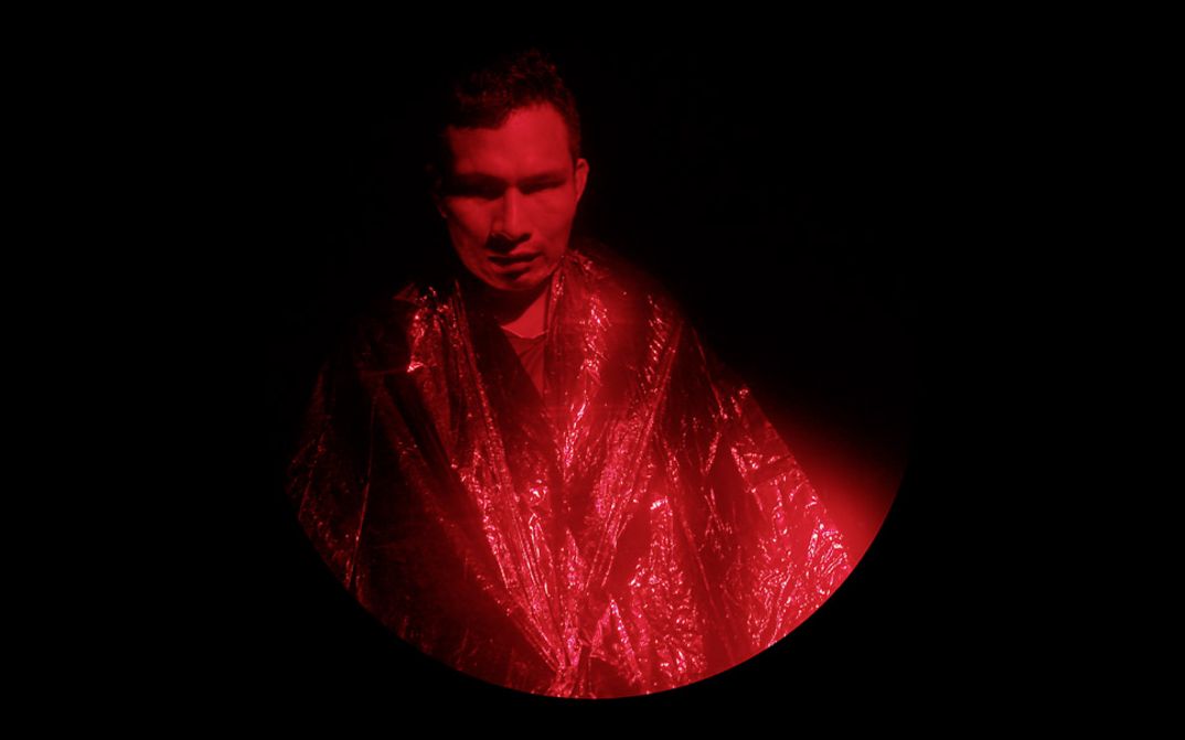 Filmstill aus dem Film "Yarokamena" von Andrés Jurado. Runder Bildausschnitt. Ein Mann mit einem silbernen Umhang wird von rotem Licht angeleuchtet.
