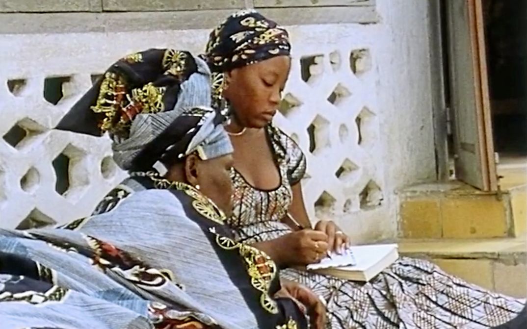 Still from the film „Man sa yay“ by Safi Faye. Two women are sitting against the wall of a house. The woman on the right is writing something on a piece of paper, while the woman on the left is looking at the paper.