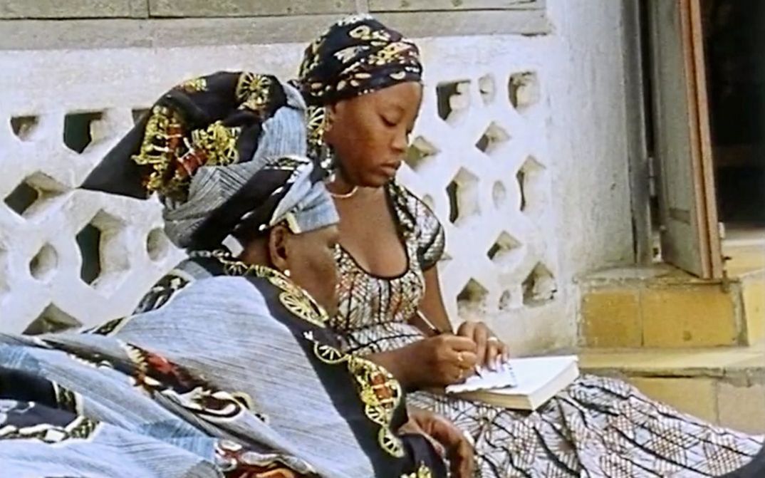 Filmstill aus „Man sa yay“ von Safe Faye. Zwei Frauen sitzen an einer Hauswand. Die Frau auf der rechten Seite schreibt etwas auf ein Papier, während die Frau auf der linken Seite auf das Papier schaut.
