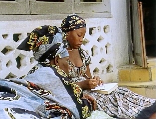 Still from the film „Man sa yay“ by Safi Faye. Two women are sitting against the wall of a house. The woman on the right is writing something on a piece of paper, while the woman on the left is looking at the paper.