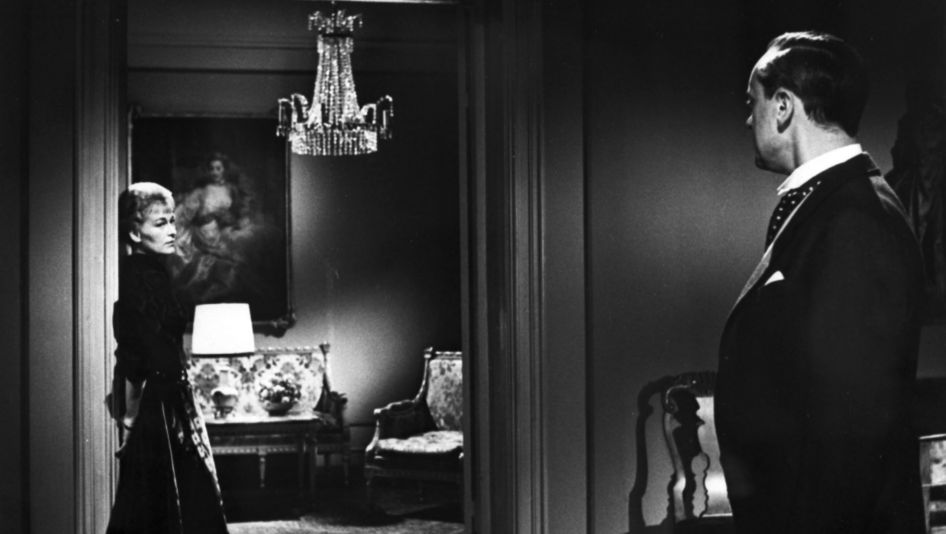 Film still from GERTRUD: In a home with a traditional, cozy decor, an elegantly dressed woman is leaning against a doorframe and looking at a man in a suit.