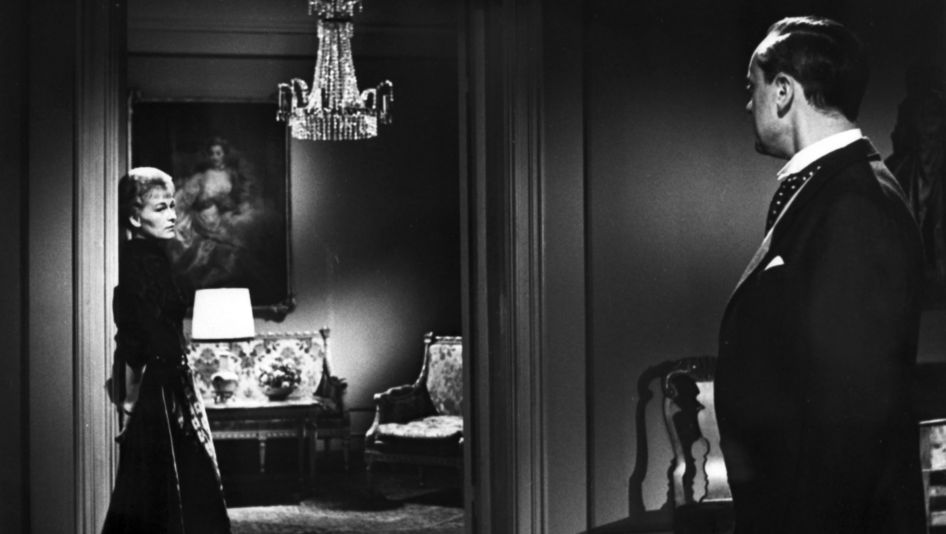 Filmstill aus GERTRUD: In einer gutbürgerlich eingerichteten Wohnung lehnt eine elegant angezogene Frau an einen Türrahmen und blickt zu einem Mann im Anzug.