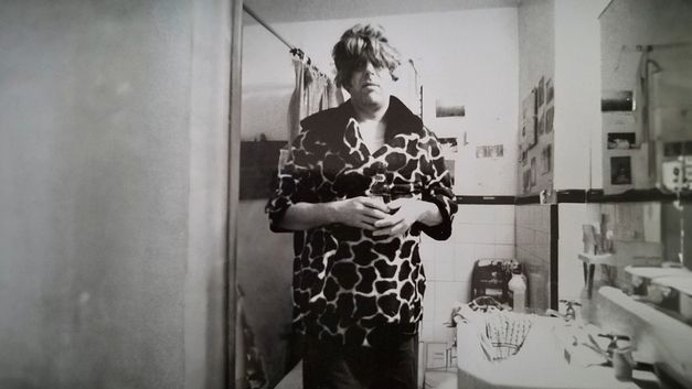 Schwarzweißaufnahme eines Mannes im Badezimmer, frontal in die Kamera schauend. Er trägt eine Perücke und eine Jacke mit Animal-Print.