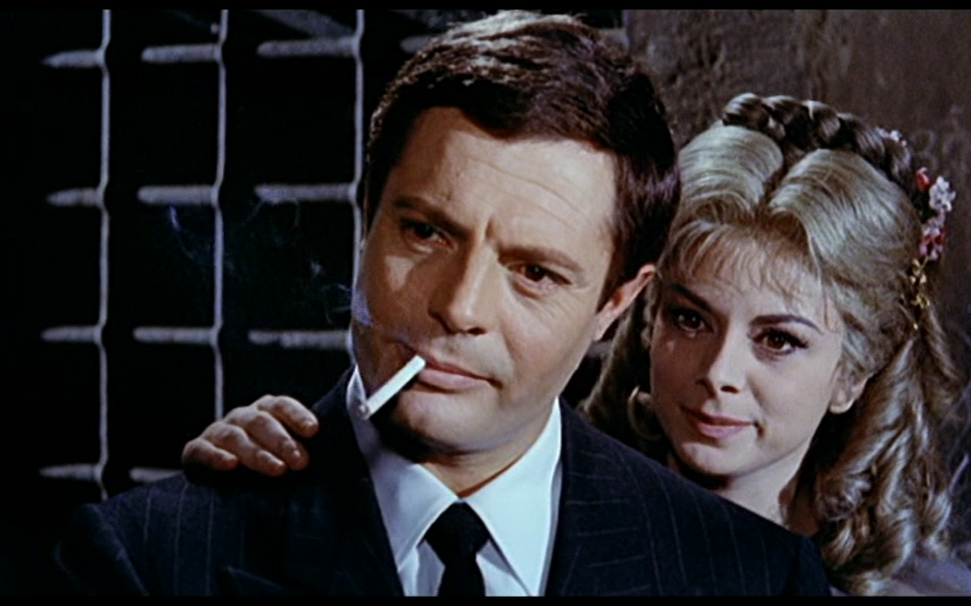 Film still from FANTASMI A ROMA: A man with a cigarette in his mouth. A woman stands behind him and puts a hand on his shoulder.