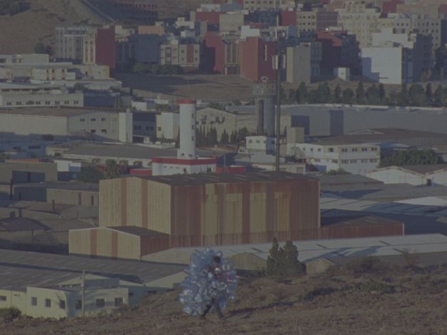 Filmstill aus dem Film „In Praise of Slowness“ von Hicham Gardaf. Eine Stadt/Kleinstadtlandschaft mit Wohnblöcken im Hintergrund, während eine Person, die eine Muschel aus leeren Plastikflaschen um sich herum hält, im Vordergrund auf einem staubigen Weg geht.