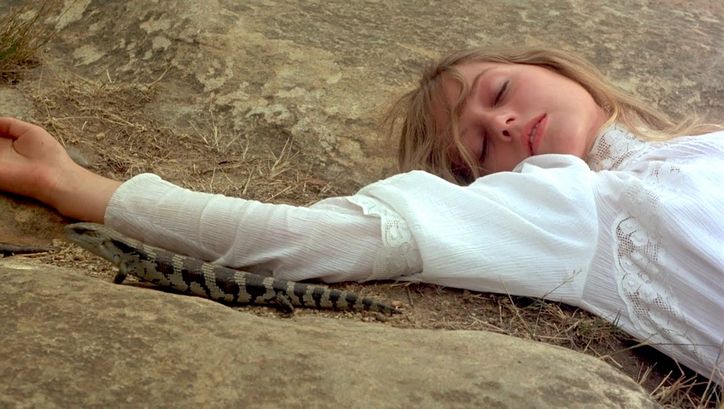 Film still from PICNIC AT HANGING ROCK: A woman lies on a rocky floor with her eyes closed. She is wearing a white dress. A lizard sits next to her arm.