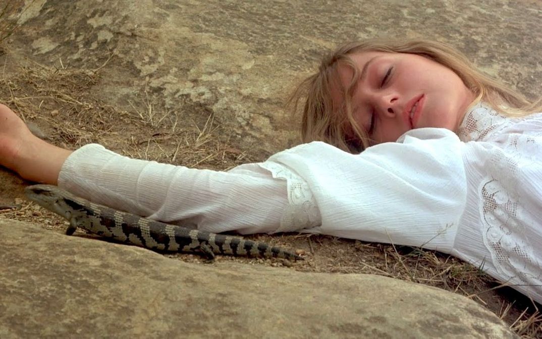 Filmstill aus PICNIC AT HANGING ROCK: Eine Frau liegt mit geschlossenen Augen auf einem Felsboden. Sie trägt ein weißes Kleid. Neben ihrem Arm sitzt eine Eidechse.