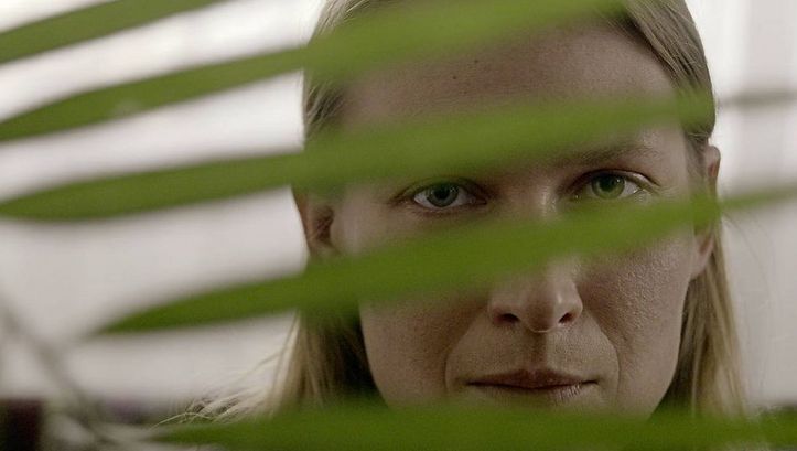Still from the film „We Haven’t Lost Our Way“  by Anka and Wilhelm Sasnal. A woman peeks through the leaf of a plant. 