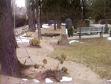 Still from the film "Normality 1–X" by Hito Steyerl. A cementary with a tombstone with an inscripition in Hebrew in the background. A man is standing in front of it. 