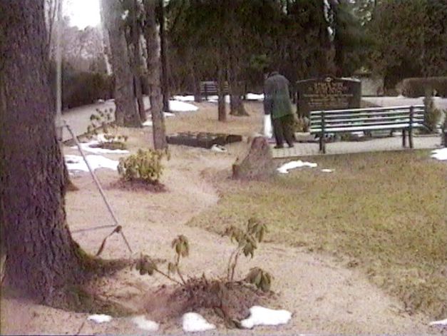 Filmstill aus „Normalität 1–10“ von Hito Steyerl. Eine Friedhofsszene. Im Hintergrund ein Grabstein mit hebräischer Schrift, vor dem ein Mann steht, der gerade im Gehen begriffen ist.