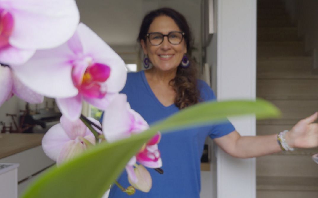 Frau steht lächelnd in einer Wohnungstür, im Vordergrund sind unscharf Blüten einer Pflanze zu sehen.
