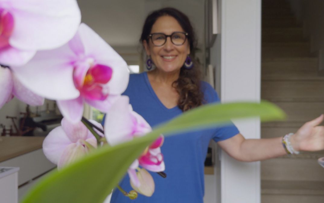 A woman smiles while standing in a doorway, with blurred flower blossoms visible in the foreground.