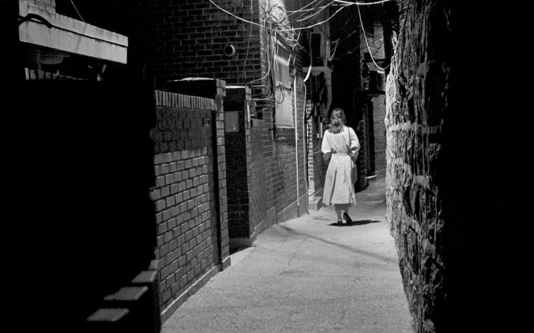 Black and white image of a woman, with her back to the camera, walking by night through a narrow alley.