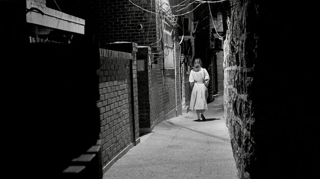 Black and white image of a woman, with her back to the camera, walking by night through a narrow alley.