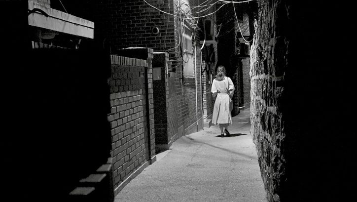 Black and white image of a woman, with her back to the camera, walking by night through a narrow alley.