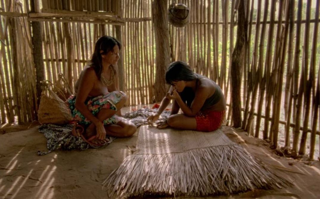 Film still from CHUVA É CANTORIA NA ALDEIA DOS MORTOS: Two indigenous Krahô sit on the floor in a hut.