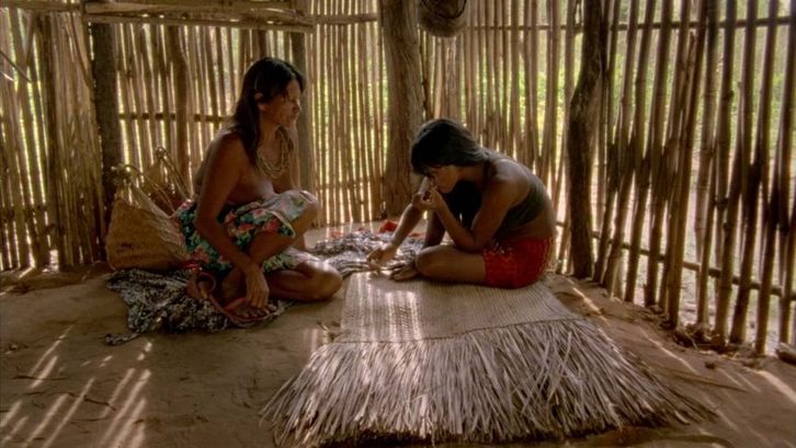 Film still from CHUVA É CANTORIA NA ALDEIA DOS MORTOS: Two indigenous Krahô sit on the floor in a hut.