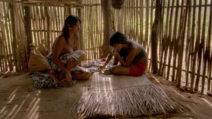 Film still from CHUVA É CANTORIA NA ALDEIA DOS MORTOS: Two indigenous Krahô sit on the floor in a hut.