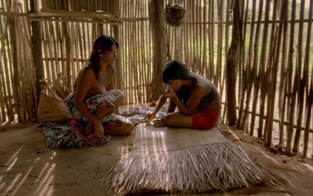 Film still from CHUVA É CANTORIA NA ALDEIA DOS MORTOS: Two indigenous Krahô sit on the floor in a hut.