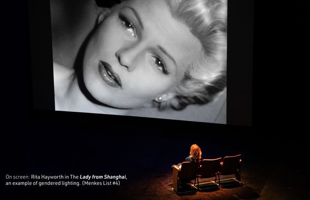 Film still from “Brainwashed“. On a dark stage, a woman sits with the back turned to us. She is looking at a bid screen. On it, we see a black and white image, a close-up of the face of Rita Hayworth.