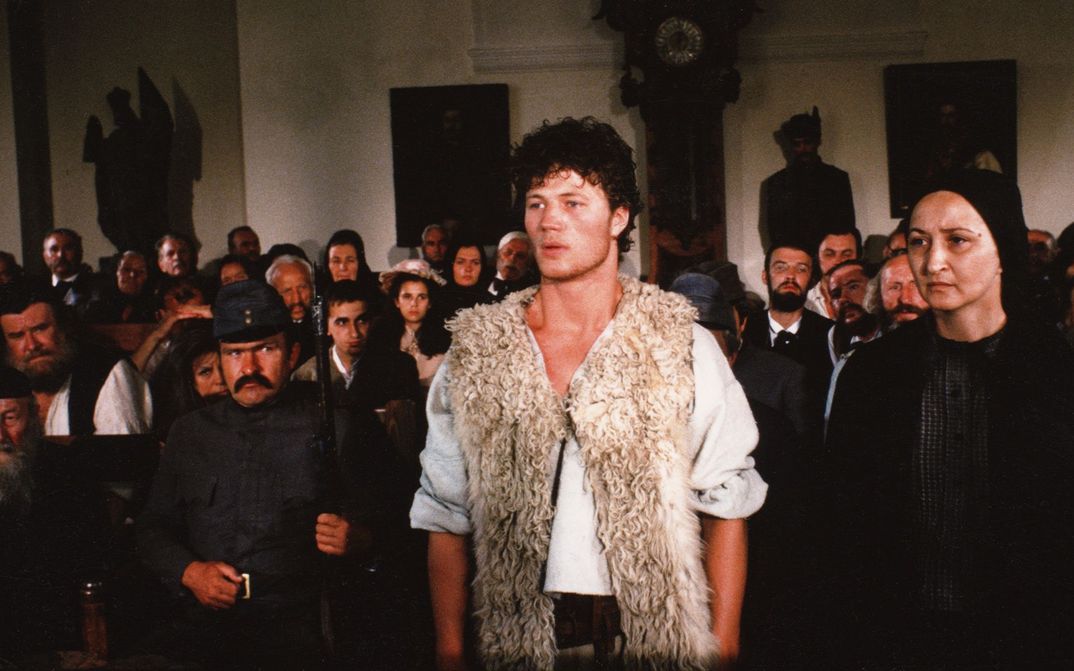 Filmstill aus MEMORIES OF A RIVER: Menschen stehen und sitzen dichtgedrängt in einem Raum. In der Mitte steht ein junger Mann mit einer Fellweste.