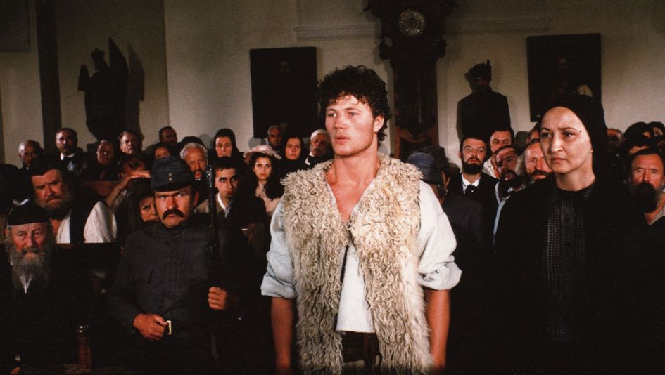 Film still from MEMORIES OF A RIVER: ople are standing and sitting crowded together in a room. In the middle stands a young man wearing a fur vest.