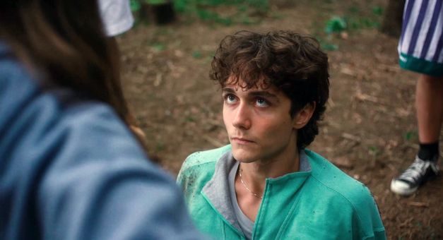 Junger Mann liegt auf erdigem Boden und blickt zu stehender Person auf.