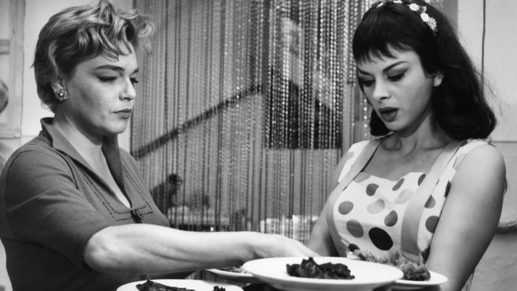 Film still from ADUA E LE COMPAGNE: Two women are standing in a restaurant, one of them is carrying four plates.