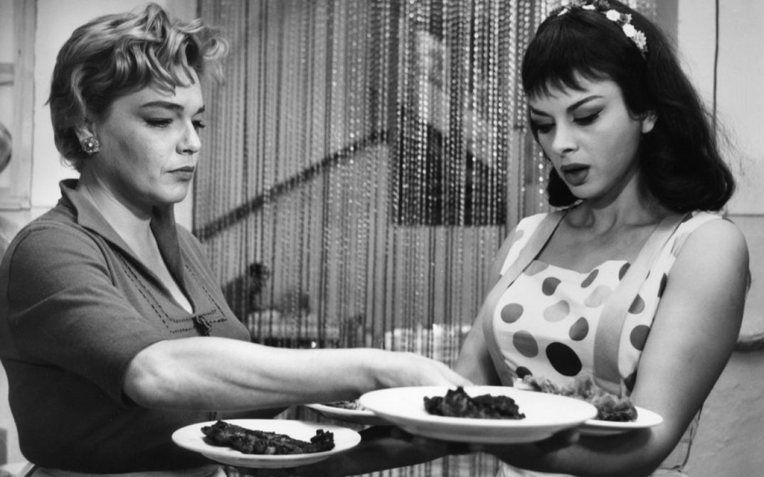 Film still from ADUA E LE COMPAGNE: Two women are standing in a restaurant, one of them is carrying four plates.
