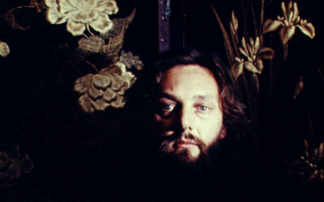 Film still from EMILY – THIRD PARTY SPECULATION: Against a dark background, a man with dark hair and a beard looks into the camera.