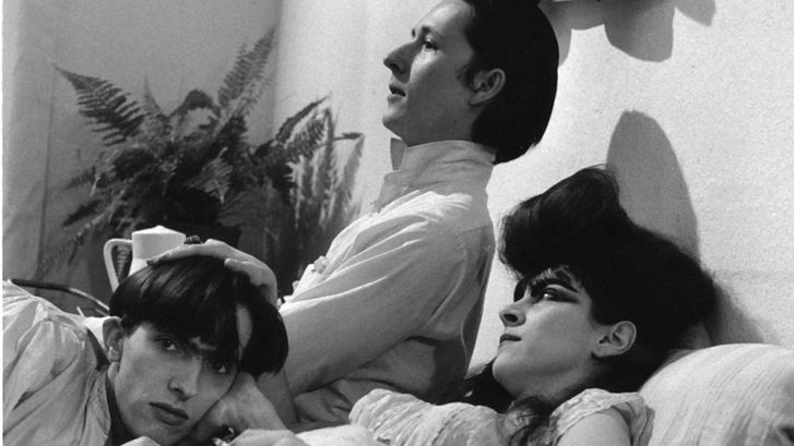 Film still from TAGEDIEBE: Three young people—two men and a woman with a striking hairstyle—are lying fully clothed in a bed.